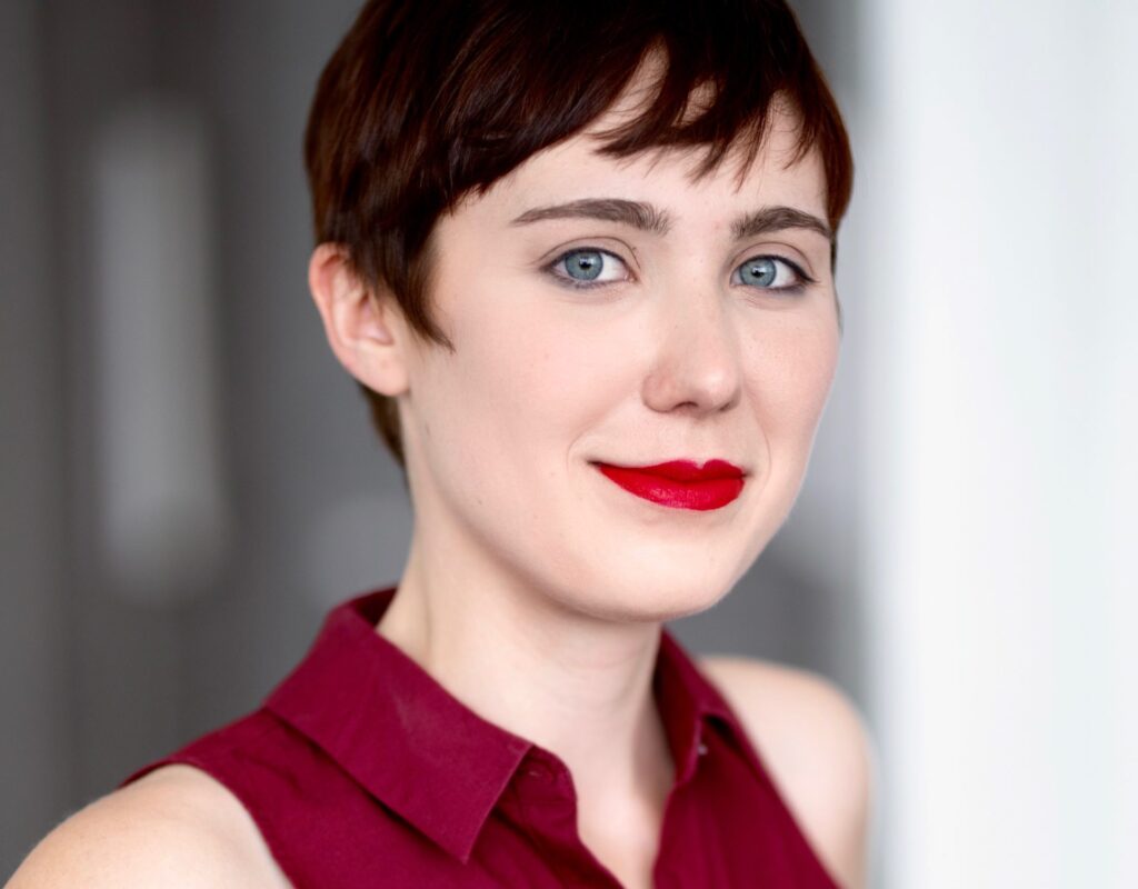 Photo of a blue-eyed white woman with a dark red pixie cut and red lipstick in a burgundy sleeveless collared shirt.